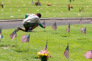 A youth kneels while placing a small U.S. flag at a grave amid rows of similar flags at other graves.