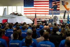 A man wearing business attire stands next to an aircraft on a stage and talks to an audience; behind him, an American flag hangs in the background.