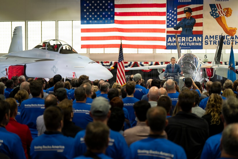 A man wearing business attire stands next to an aircraft on a stage and talks to an audience; behind him, an American flag hangs in the background.