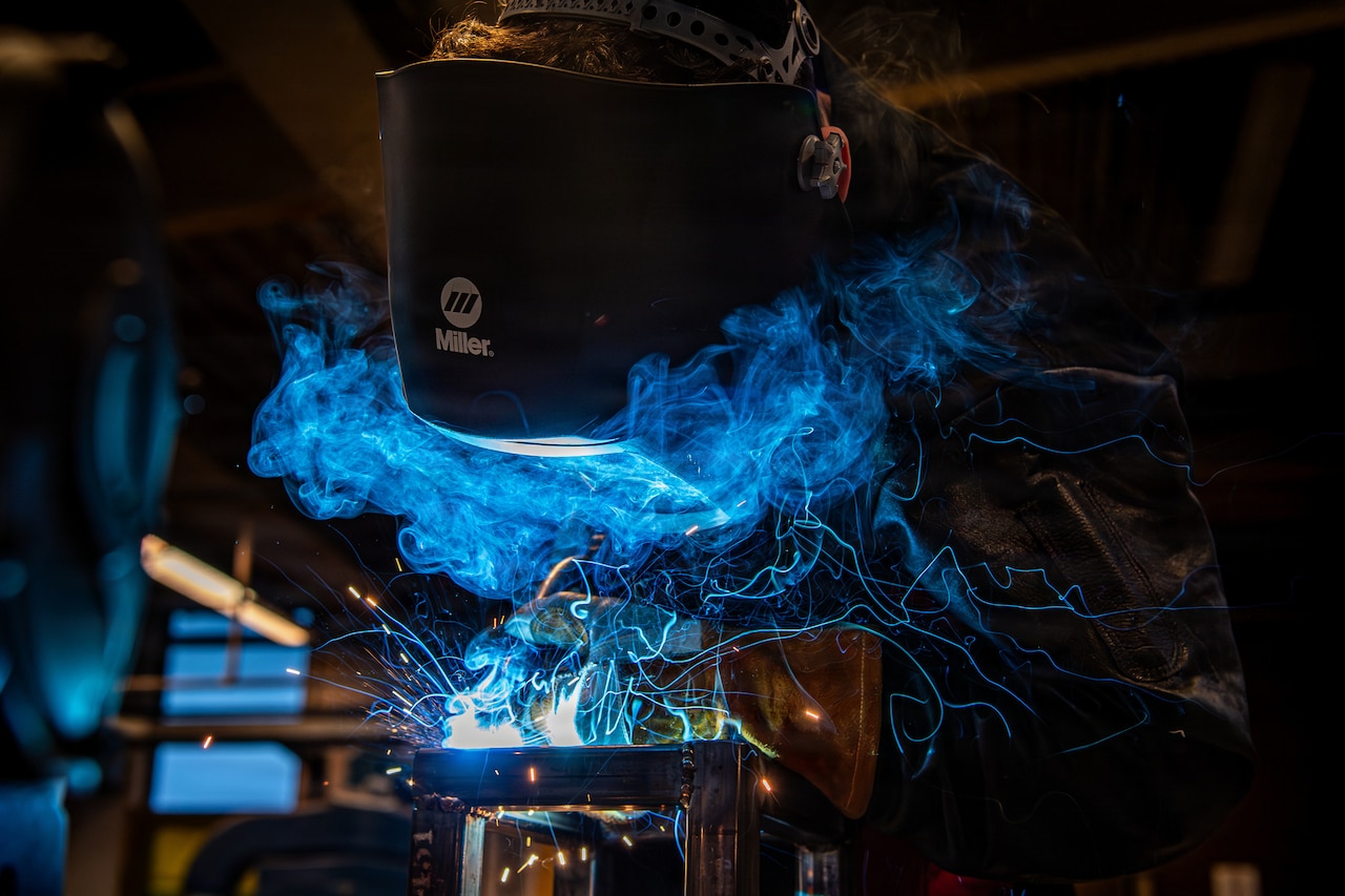 A person wearing a welding face shield bends over to use a tool to fuse metal, as blue sparks and smoke emit from the reaction.