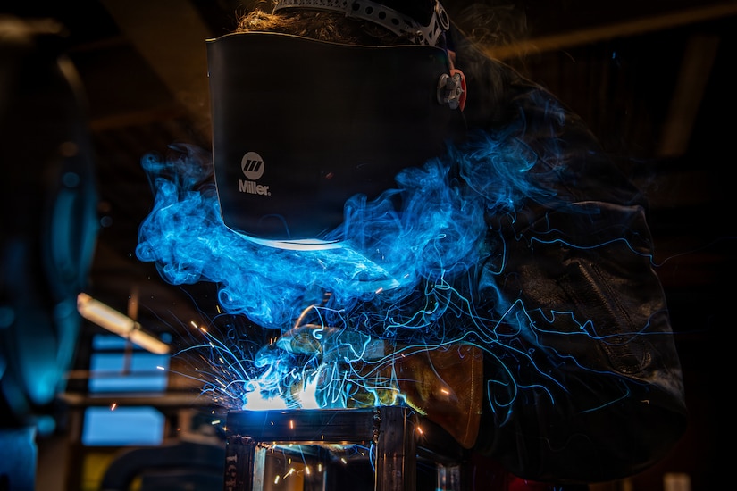 A person wearing a welding face shield bends over to use a tool to fuse metal, as blue sparks and smoke emit from the reaction.