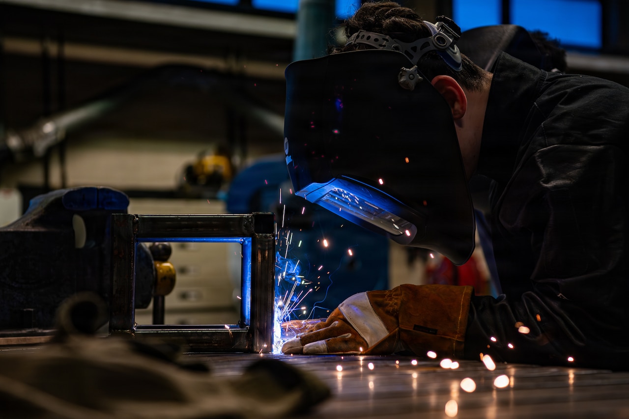 A person wearing a welding face shield and gloves bends over a table and uses a tool to fuse metal, as blue sparks emit from the reaction.
