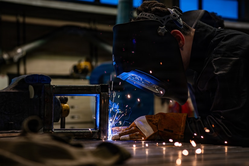 A person wearing a welding face shield and gloves bends over a table and uses a tool to fuse metal, as blue sparks emit from the reaction.