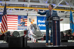 A man in a business suit stands behind a lectern, gesturing with one hand as he speaks, with a jet, American flags and other signage in the background.