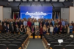 Large group posing for photo on stage with a blue screen in back and flags on both sides