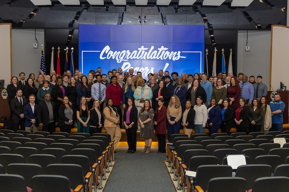 Large group posing for photo on stage with a blue screen in back and flags on both sides