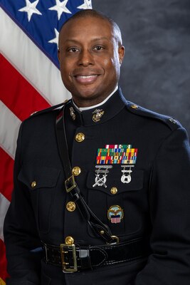 A dark-skinned man in a dress uniform for the Marine Corps. There is a flag behind him.
