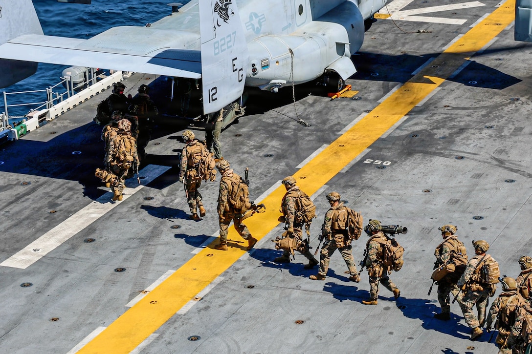 Marines seen from above walk in a line to the open back of a parked aircraft on the deck of a ship at sea.