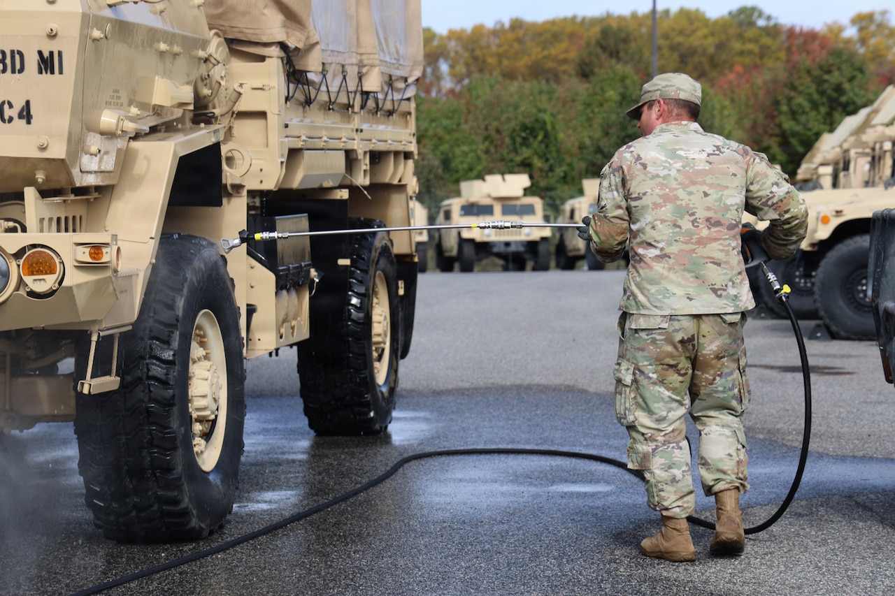 A man in a camouflage military uniform uses a tool attached to a hose to spray solution on the tire of a large military vehicle, outside in a parking lot with similar vehicles in the background and trees in the distance.