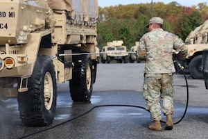 A man in a camouflage military uniform uses a tool attached to a hose to spray solution on the tire of a large military vehicle, outside in a parking lot with similar vehicles in the background and trees in the distance.