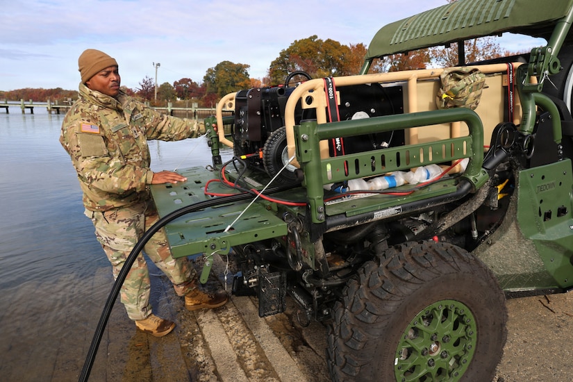 A man in a camouflage military uniform stands next to a military truck with equipment in the back. A body of water and trees are in the background.
