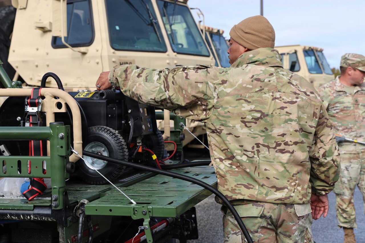 A man in a camouflage military uniform stands in a parking lot next to a military truck with equipment in the back. People in similar attire and other vehicles are in the background.