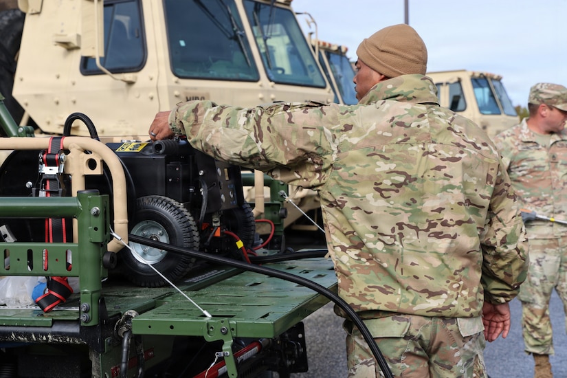 A man in a camouflage military uniform stands in a parking lot next to a military truck with equipment in the back. People in similar attire and other vehicles are in the background.