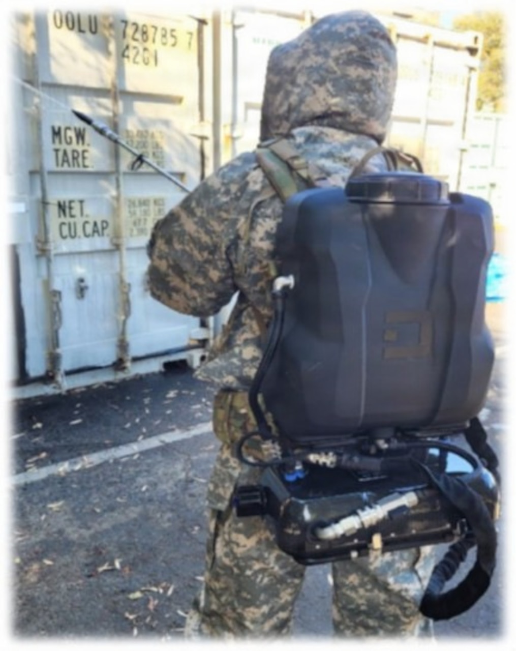 A person wearing a camouflage military uniform and a large backpack with hoses attached, uses a tool to spray solution on large military storage containers outside in a parking lot.