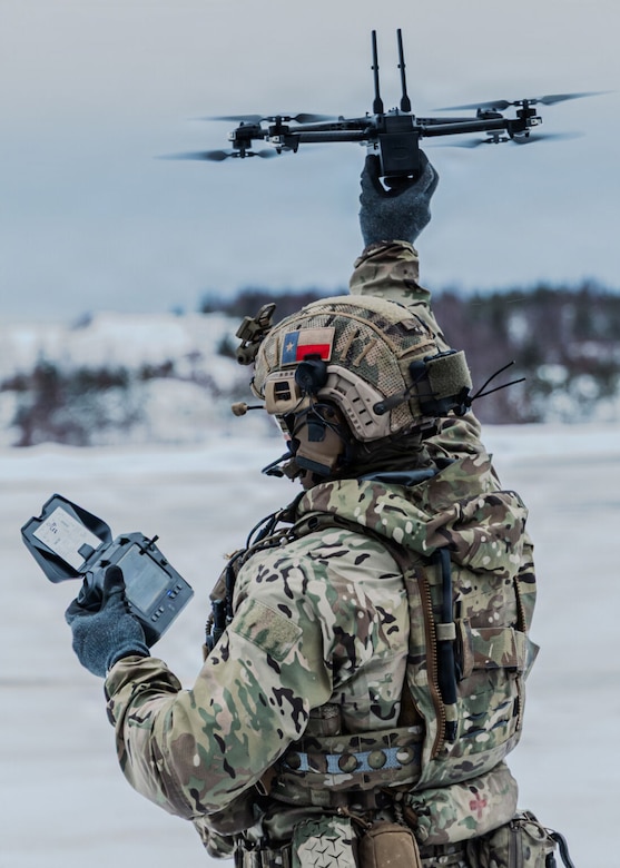 A person in a camouflage cold-weather military uniform holds a drone over his head with one hand and a remote control in the other, outside in a snowy environment with trees in the distance.