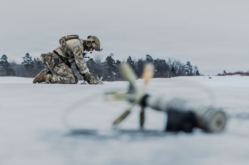 A person in a camouflage cold-weather military uniform crouches down toward a snow-covered ground to place a piece of military equipment on the ground. Another piece of military equipment is in the foreground, and trees are in the distance.