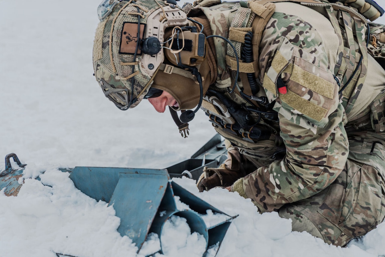 A man in a camouflage cold-weather military uniform crouches down on a snow-covered ground as he works with a piece of military equipment.
