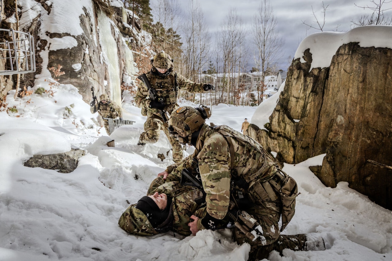 A person in a camouflage military uniform bends down to attend to another person in similar attire next to large rocks on a snow-covered ground, while two people in military uniforms  walk toward them. Trees and buildings are in the distance.