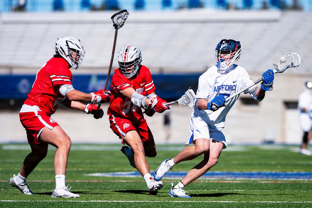 An Air Force lacrosse player runs with the ball as opposing players try to stop them during a game at a stadium on a sunny day.