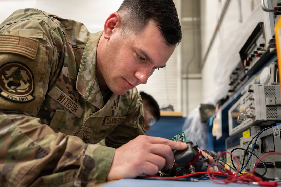 An airman leans over to inspect an actuator next to other gadgets in a room.