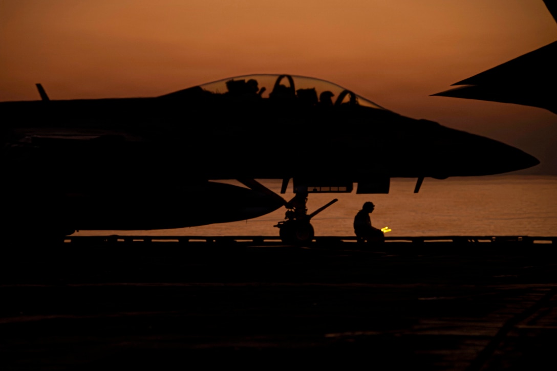 An aircraft sits on the flight deck of a ship under an orangish sky as seen in silhouette.