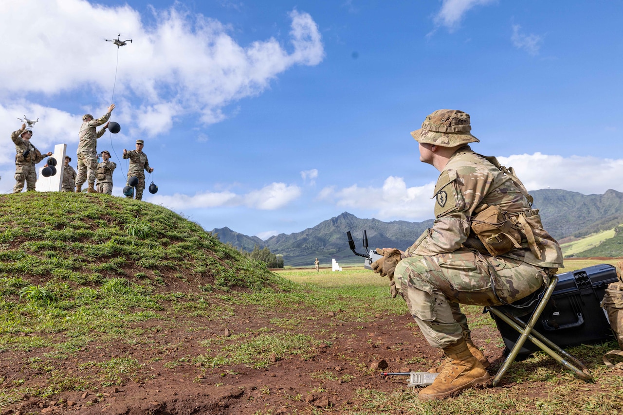 A sitting soldier uses a remote control as fellow soldiers standing on a hill launch drones into a sunny, blue sky with mountains in the background.