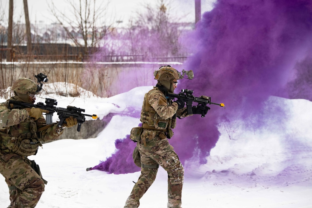 Two soldiers wearing tactical gear aim weapons while moving through snow as clouds of purple smoke fill the air.
