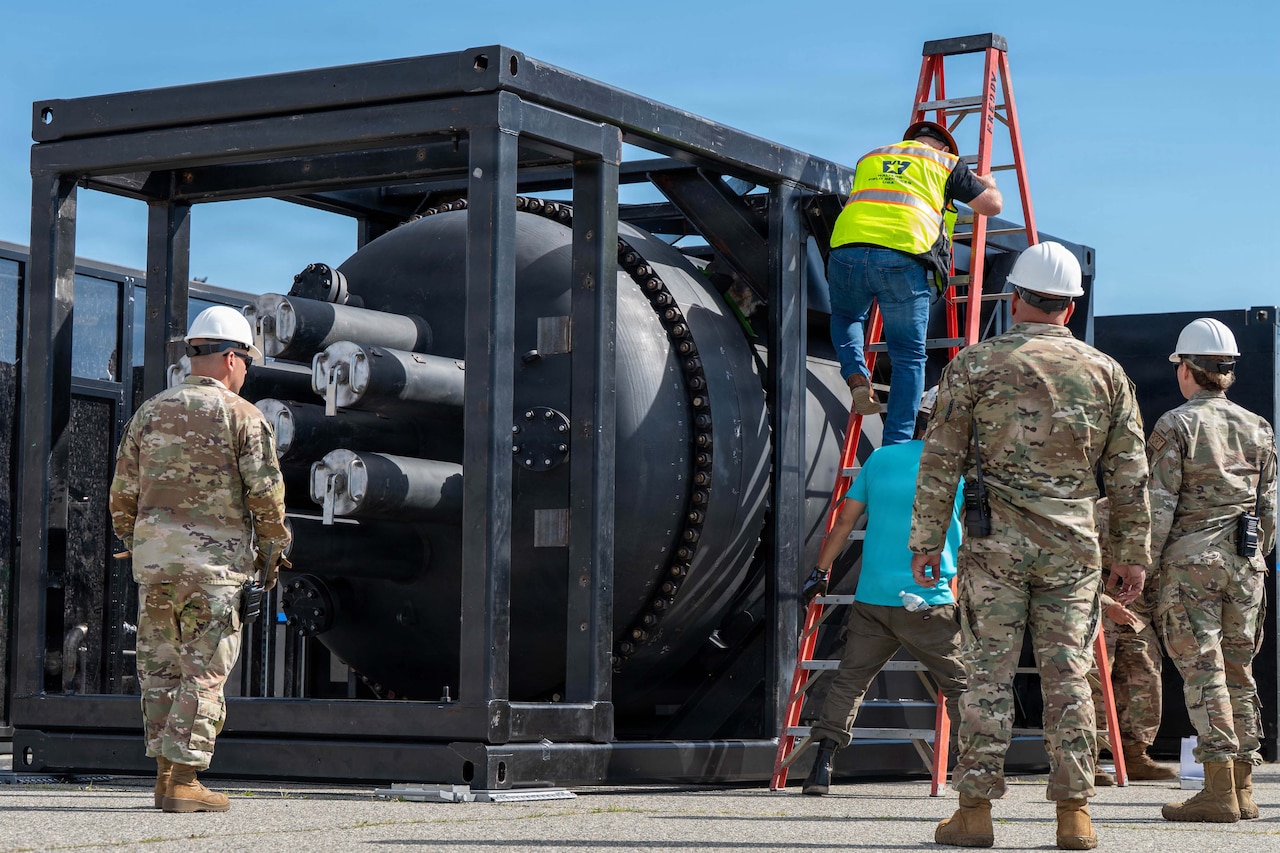 Four people wearing camouflage military uniforms and hard hats stand around a metal cage with a large tub inside, as a man in a yellow vest climbs a ladder near the cage.