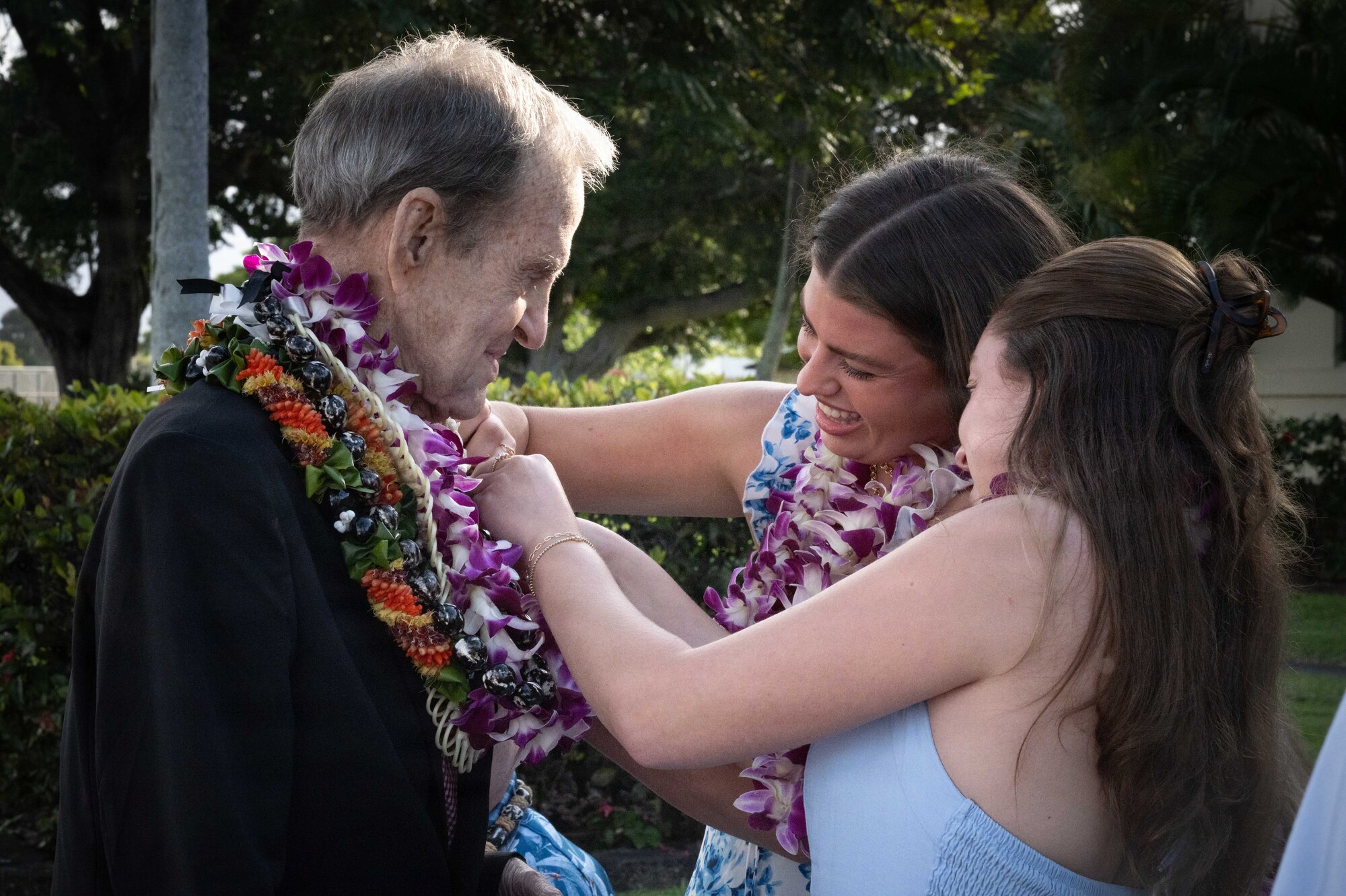 An elderly man wearing Hawaiian lei's stands as two of his granddaughters put a pin on his suit.
