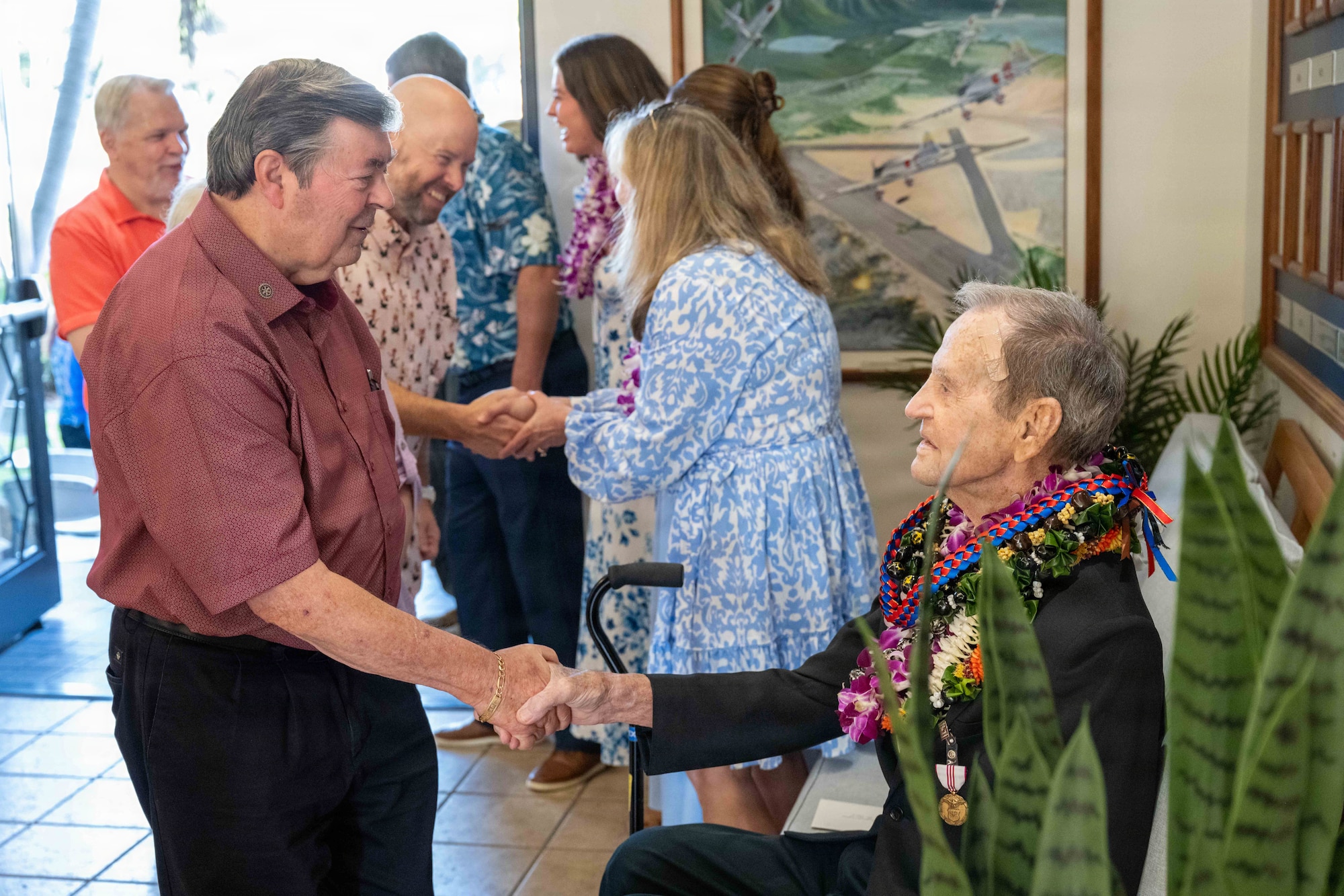 Elderly man sits as another man shakes his hand.