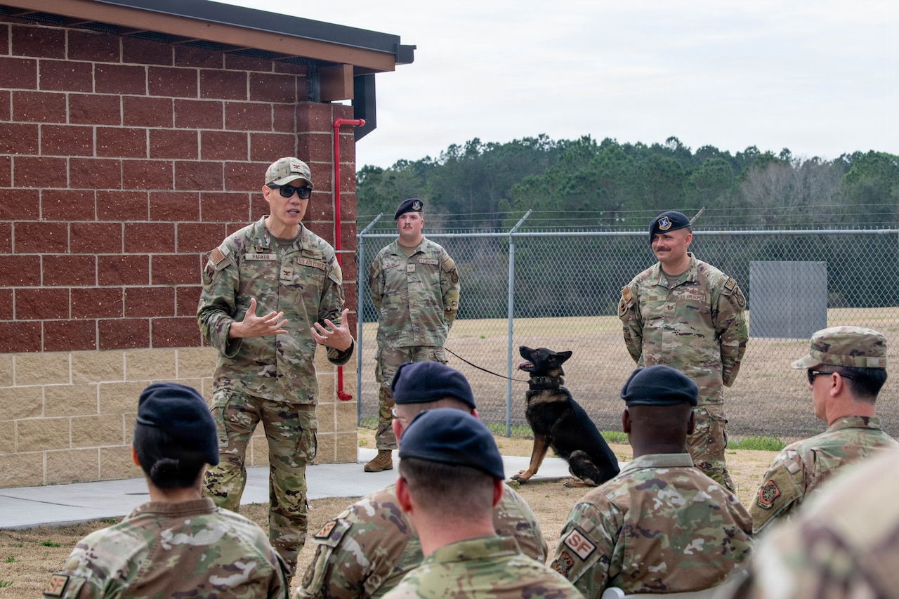 A man in a camouflage military uniform and sunglasses speaks to a seated group of people in similar attire. Behind the man is a building and two other men in camouflage military uniforms standing with their arms behind their backs; one of the men is holding the leash of a dog.