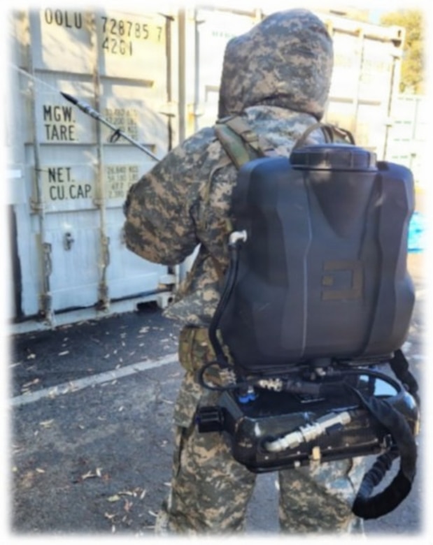 A person wearing a camouflage military uniform and a large backpack with hoses attached, uses a tool to spray solution on large military storage containers outside in a parking lot.