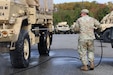 A man in a camouflage military uniform uses a tool attached to a hose to spray solution on the tire of a large military vehicle, outside in a parking lot with similar vehicles in the background and trees in the distance.