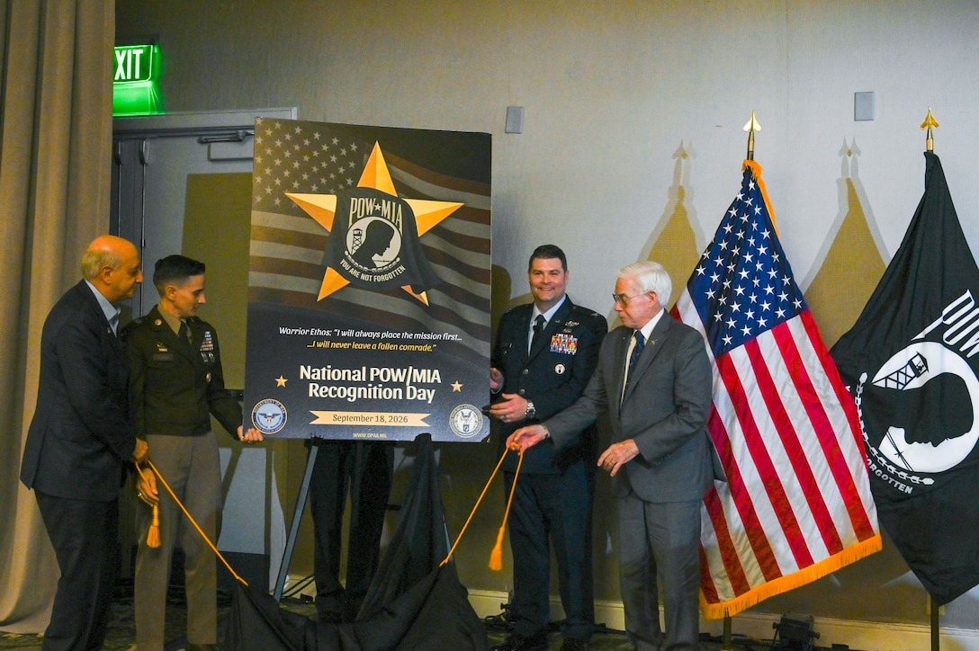 The Defense POW/MIA Accounting Agency Senior Enlisted Advisor, U.S. Army Sgt. Maj. Lauren Flores, and DPAA Chief of Staff U.S. Air Force Col. Derek Rankin stand alongside two former prisoners of the Vietnam War during the unveiling of the National POW/MIA Recognition Day poster at the Charleston Family Member Update, Feb. 21, 2026.