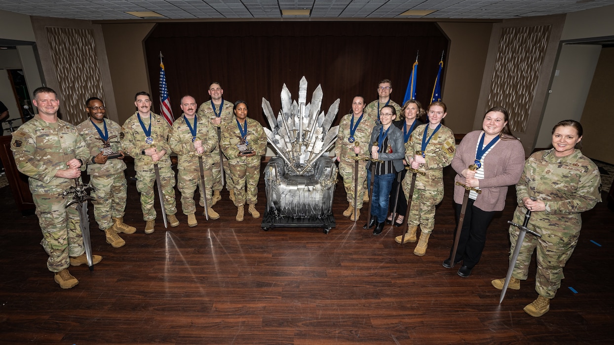 A group photo of military members, wearing camouflage uniforms, and civilians around a throne made of carboard swords