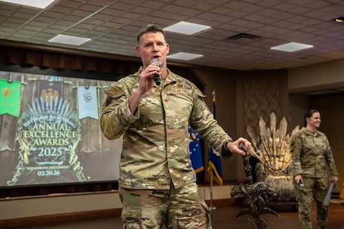 A man, wearing a camouflage military uniform, speaks into a microphone while holding a fantasy sword