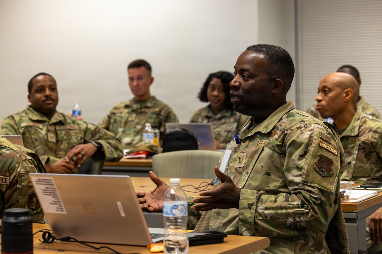 Six people wearing camouflage military uniforms sit at desks in a room with laptops in front of them; the man in the foreground is talking and gesturing with his hands.