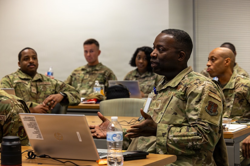 Six people wearing camouflage military uniforms sit at desks in a room with laptops in front of them; the man in the foreground is talking and gesturing with his hands.