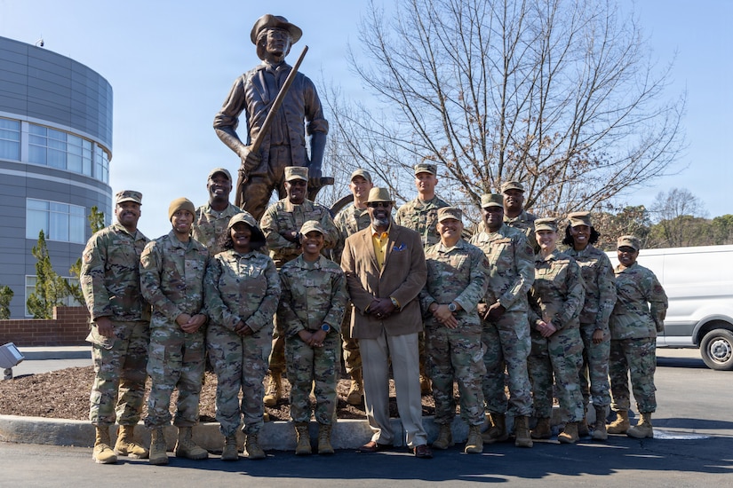 A group of men and women wearing camouflage military uniforms pose for a photo with a man in business attire in the center. Behind them is a statue of a man, and in the background are trees and an office building.