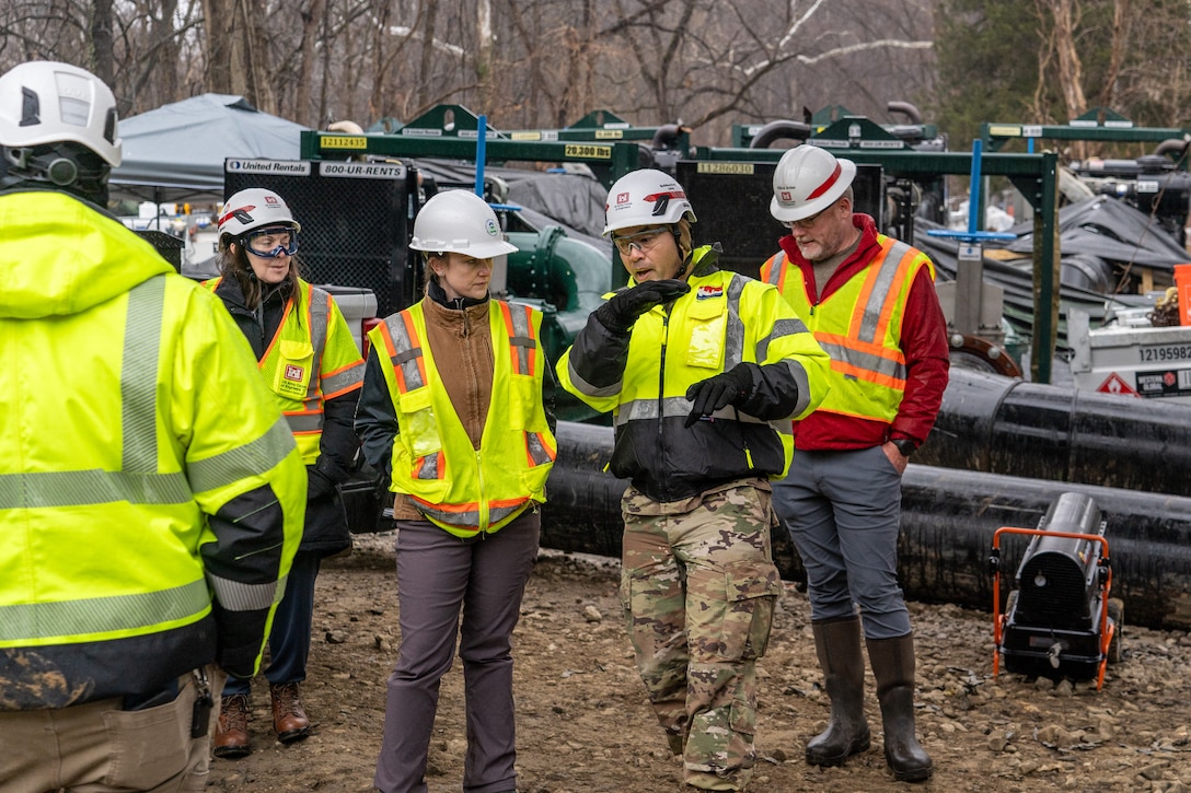 Individuals in high-visibility construction vests and jackets and white hard hats stand outside in a group discussion. They are surrounded by pipes and other response & construction equipment.