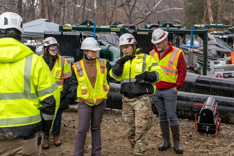 Individuals in high-visibility construction vests and jackets and white hard hats stand outside in a group discussion. They are surrounded by pipes and other response & construction equipment.
