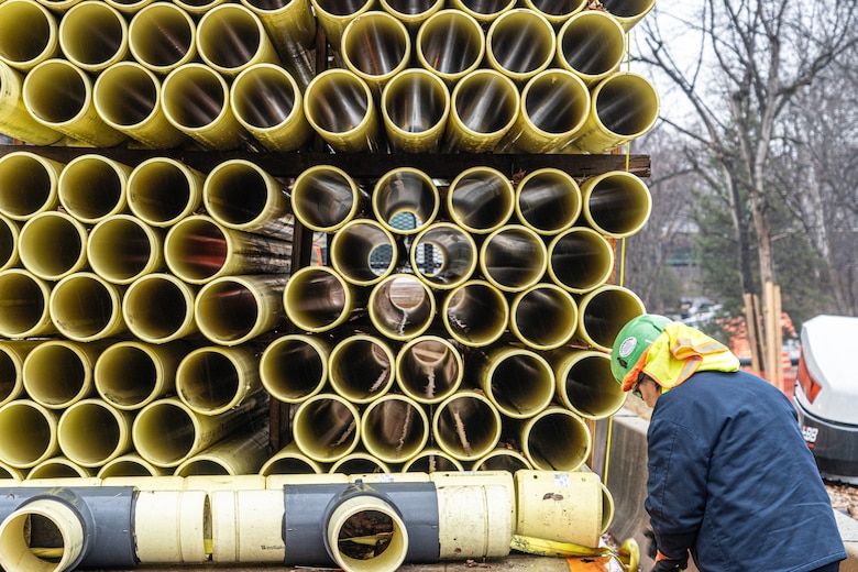 A large quantity of yellow drainage pipe is visible from one end, stacked high on a truck bed, while a worker in a green hard hat begins to remove it.