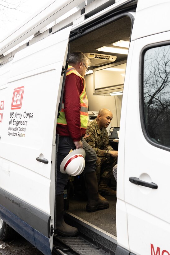 Individuals in high-visibility construction gear discuss operations from inside a white van marked "Deployable Tactical Operations System" on the side.