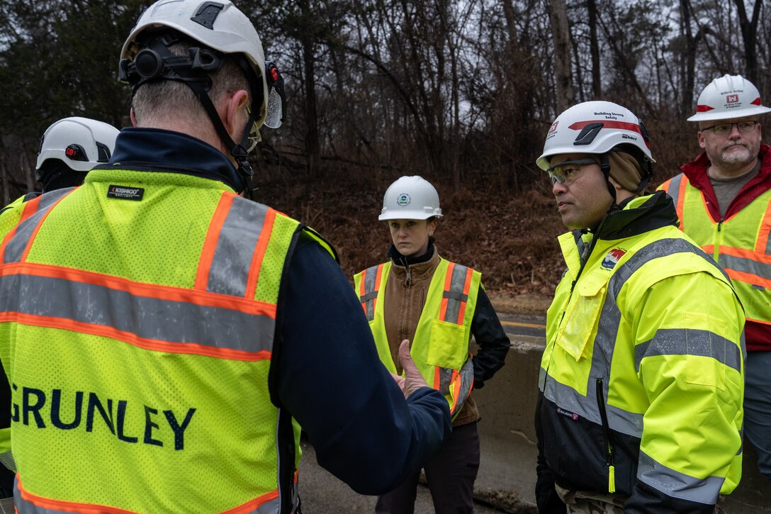 Individuals in high-visibility construction vests and jackets and white hard hats stand outside in a group discussion.