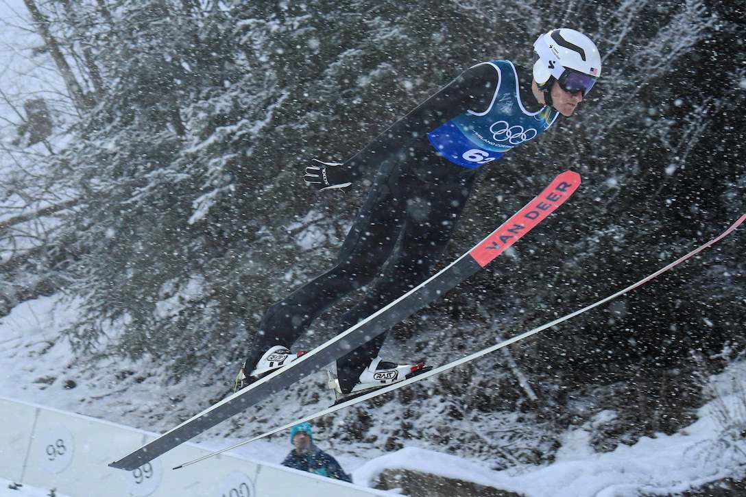 A person wearing an Olympic ski uniform, gloves, a helmet and goggles lunges forward in the air on a pair of skis in the falling snow; snow-covered trees are in the background.