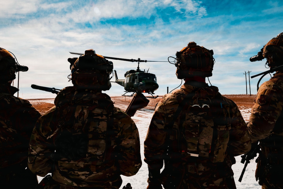 The silhouettes of four people wearing military uniforms and helmets watch a helicopter hover over a flight line with blue skies overhead.