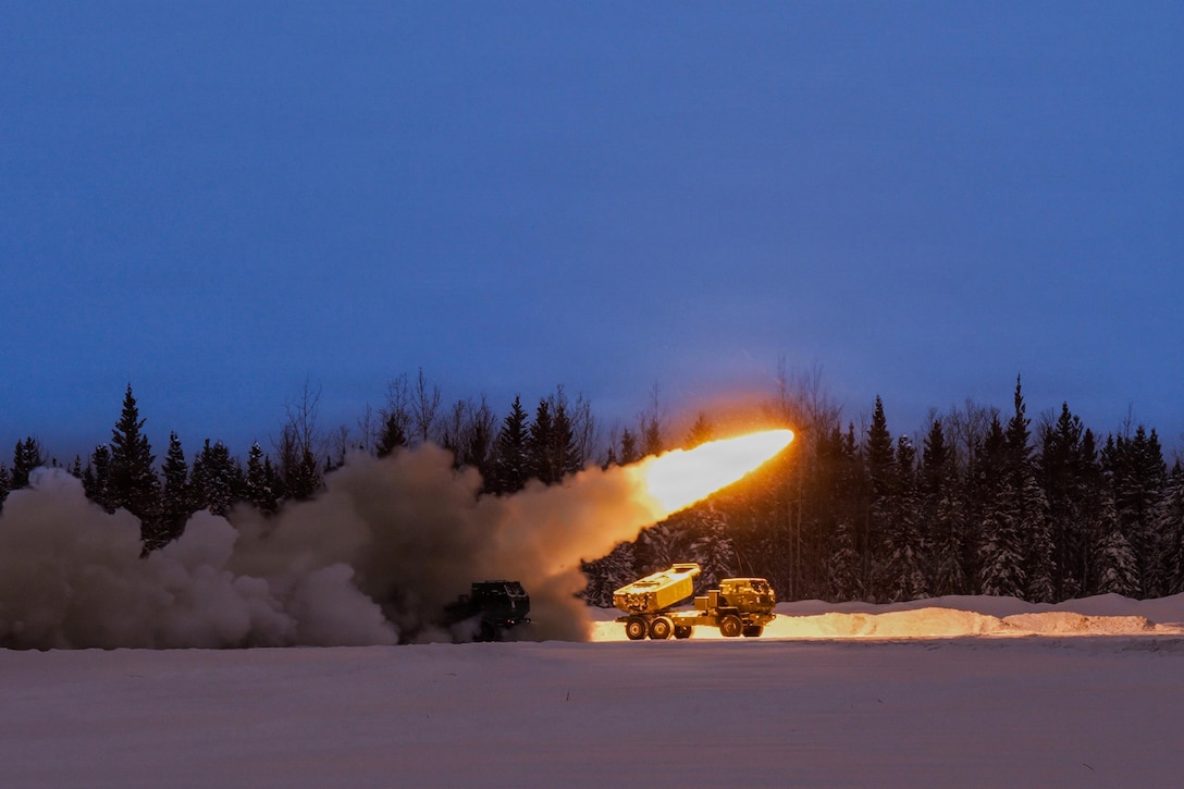 White smoke and yellow flames emit from a military vehicle parked in the snow under a dark sky, with trees in the background.