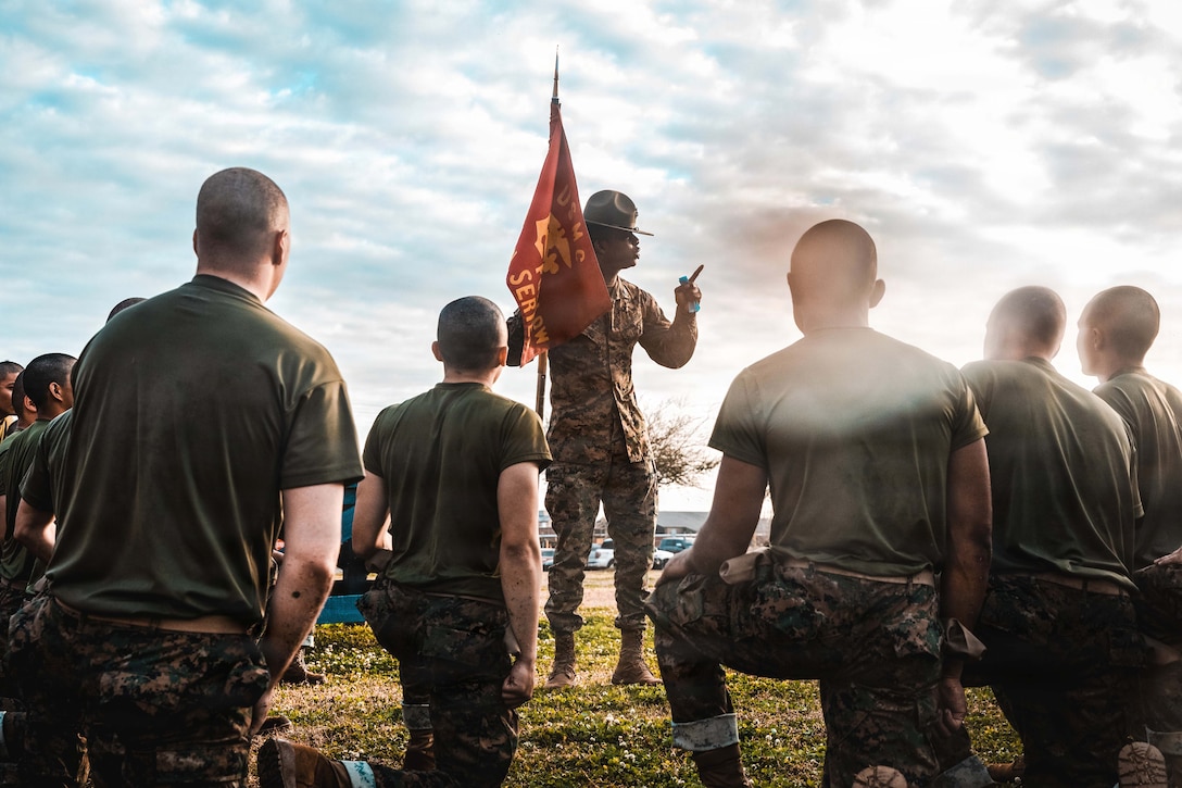 A Marine stands in the center of a field with a red flag as recruits kneel around them under a bright, cloudy sky.