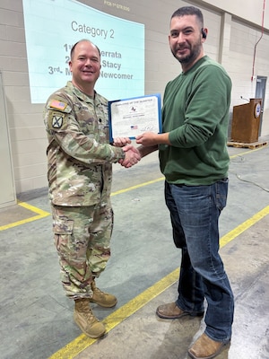 A man wearing a camouflage military uniform poses with another person holding an award certificate.