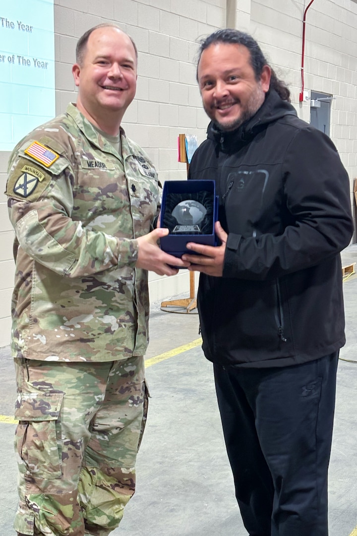 A man wearing a camouflage military uniform poses with another person holding a trophy.